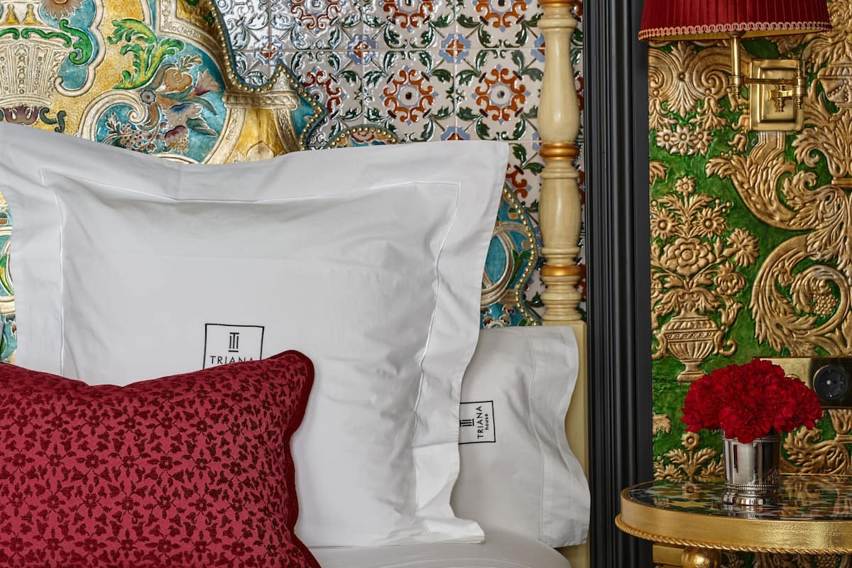 Un lit avec des oreillers blancs et rouges, un oreiller à motifs rouges, une tête de lit avec un cadre doré et une petite table sur laquelle se trouvent une lampe rouge et un vase de fleurs.