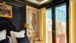 Quarto de hotel com uma cama grande, uma cabeceira, uma parede com padrão de azulejos decorativos, uma pequena mesa com um vaso e vista para um pátio através de uma porta de vidro.
