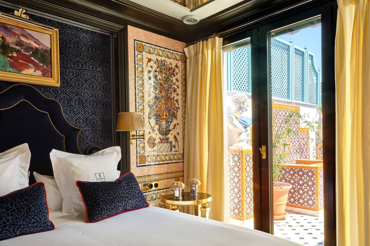 Une chambre d’hôtel avec un grand lit, une tête de lit, un mur orné d’un motif décoratif en carreaux, une petite table avec un vase, et une vue sur une cour intérieure, visible à travers une porte vitrée.