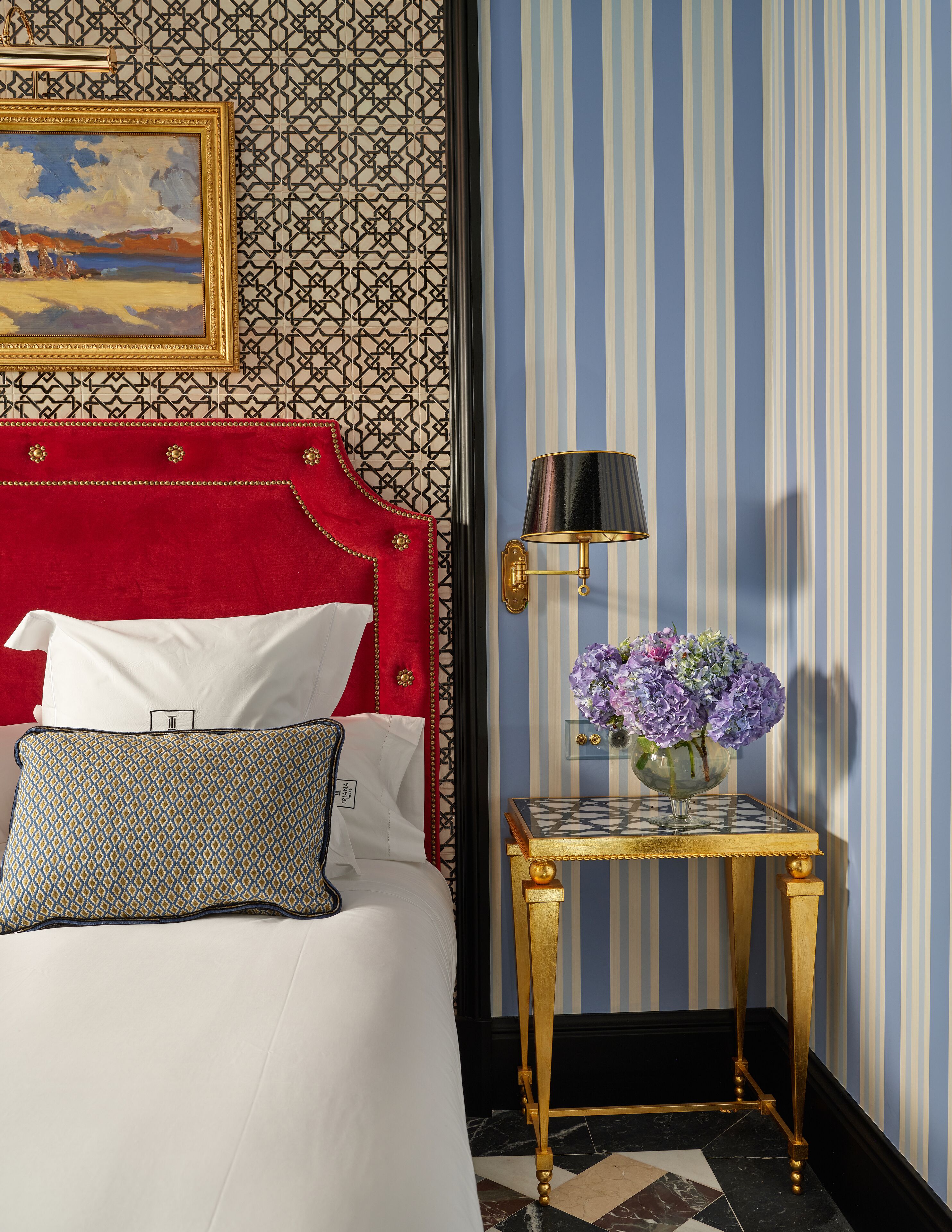 A hotel room with a red upholstered headboard, a patterned pillow, a small table with a vase of flowers, and a wall with vertical stripes.