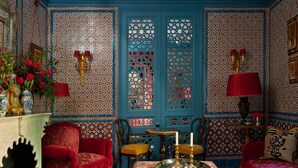 Quarto com piso xadrez, paredes estampadas em vermelho e azul, e um banquinho central com bandeja.
