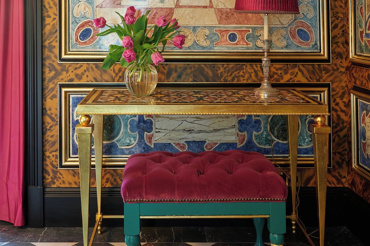 Une pièce décorée d’un tableau encadré représentant une tour et des vases, une petite table sur laquelle est posé un vase de fleurs, et un banc de couleur turquoise.