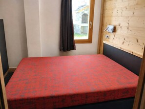 A small, single-bed room with a red patterned bedspread, a window with a curtain, and a wooden wall with a thermostat. - Renovated apartment near slopes with free parking (La Plagne Montchavin Les Coches)