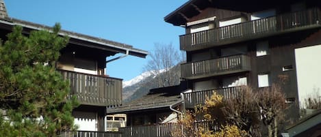 A traditional chalet-style building with multiple balconies and a wooden facade, surrounded by trees and a clear blue sky.