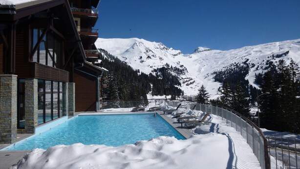 Blick auf ein Schwimmbecken neben einem Gebäude, mit blauem Wasser, umgeben von Schnee und Bergen.