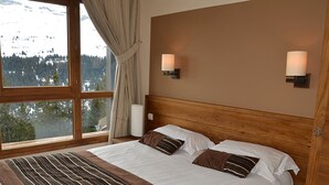 Ein Schlafzimmer mit einem großen Bett, zwei Nachttischlampen und Blick auf einen verschneiten Berg durch ein großes Fenster.