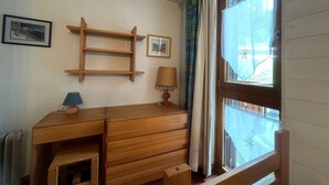 Un escriptori de fusta amb calaixos i una prestatgeria, una làmpada i una finestra amb cortines.