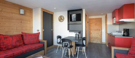 Un espacio habitable compacto con un sofá rojo, una mesa de comedor con sillas, una zona de cocina y un televisor montado en la pared.