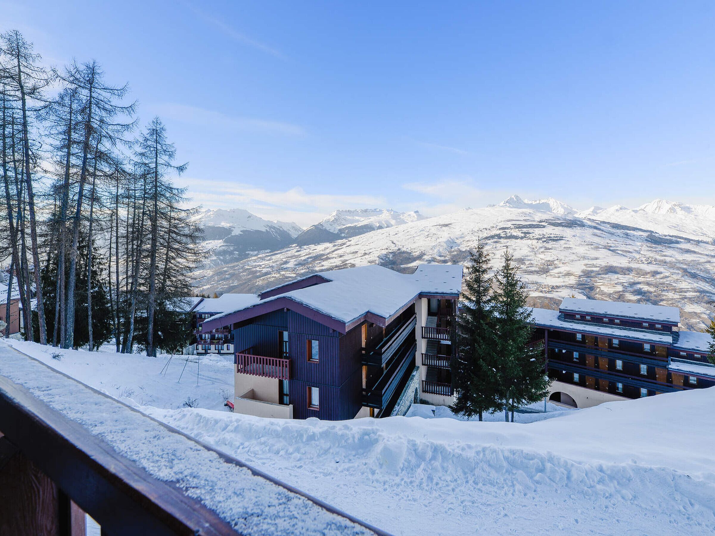Une station de montagne enneigée, dotée de bâtiments modernes, dont un immeuble à plusieurs étages avec des balcons, et où l'on peut apercevoir un télésiège.
