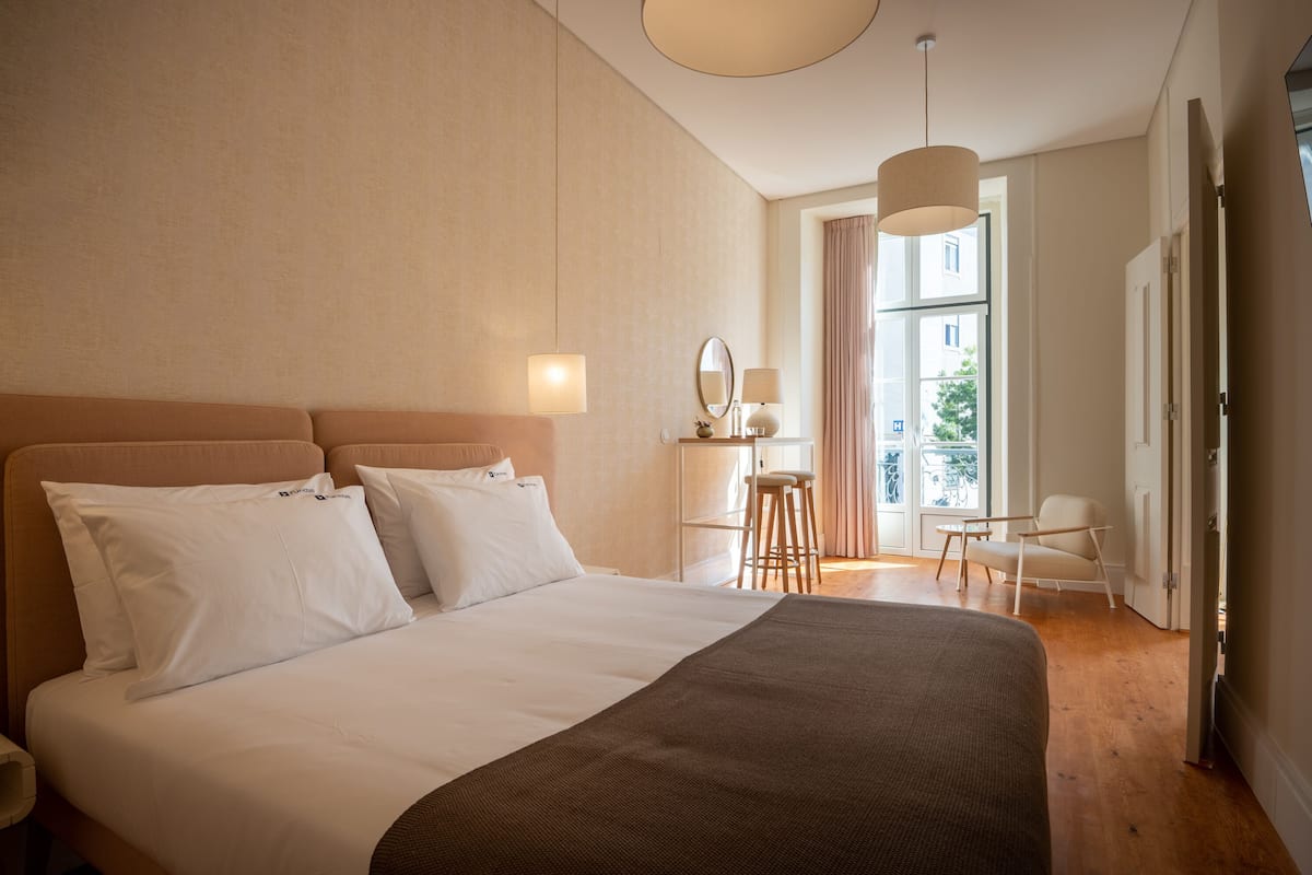Una habitación de hotel con una cama grande, una zona de estar con una silla y una mesa, un espejo y una ventana con cortinas.