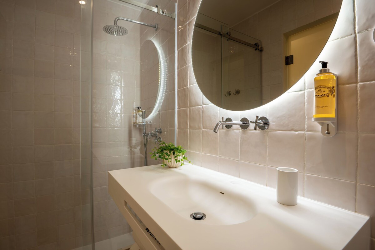 Un baño moderno con ducha, lavamanos, espejo y una botella de loción corporal Castelfelso.