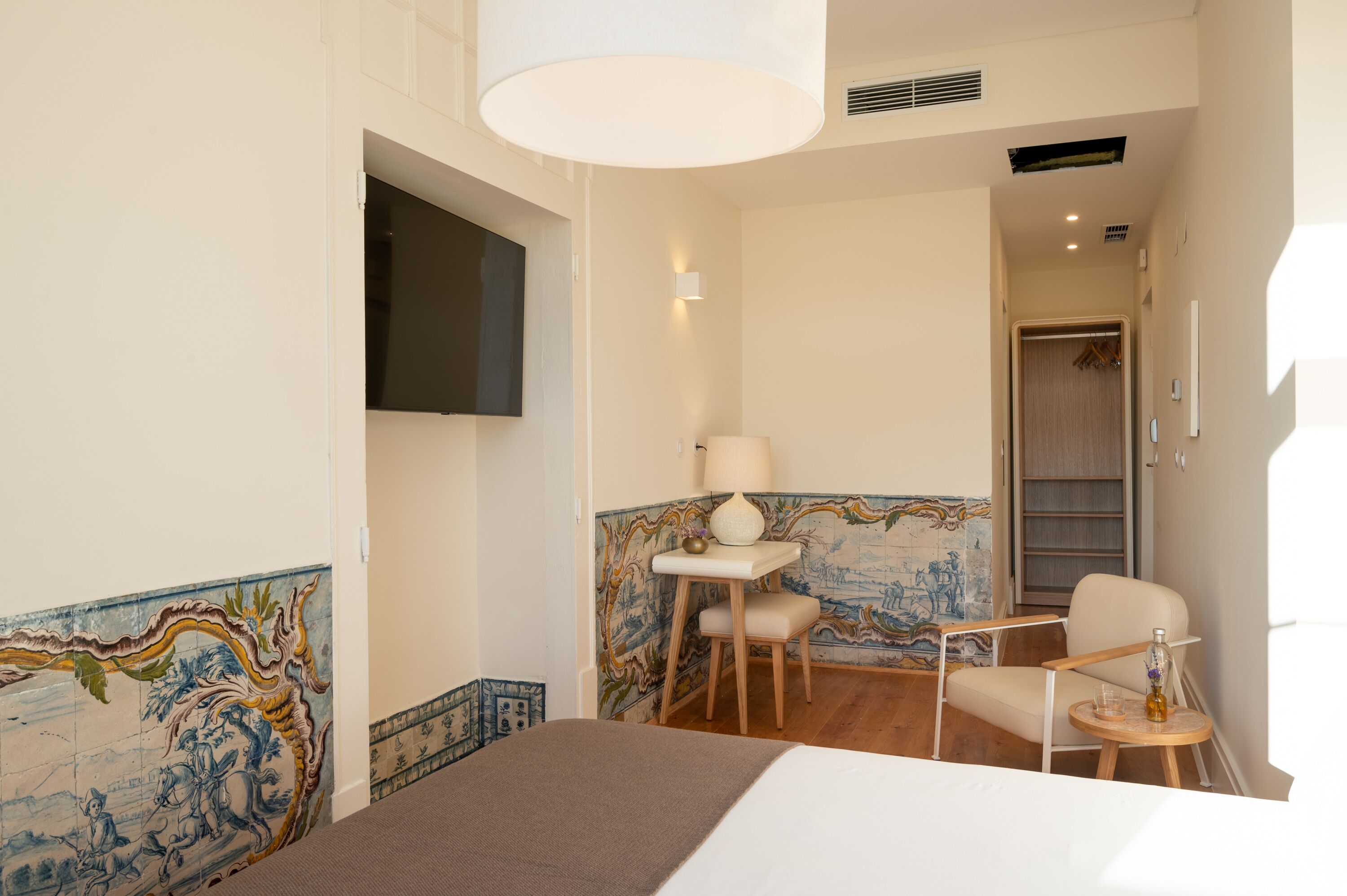 A hotel room with a bed, a small table with a lamp, a chair, and a TV mounted on the wall.