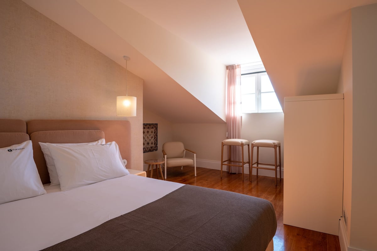 Una habitación de hotel con una cama grande, una silla, una mesita y un ventanal con cortinas.