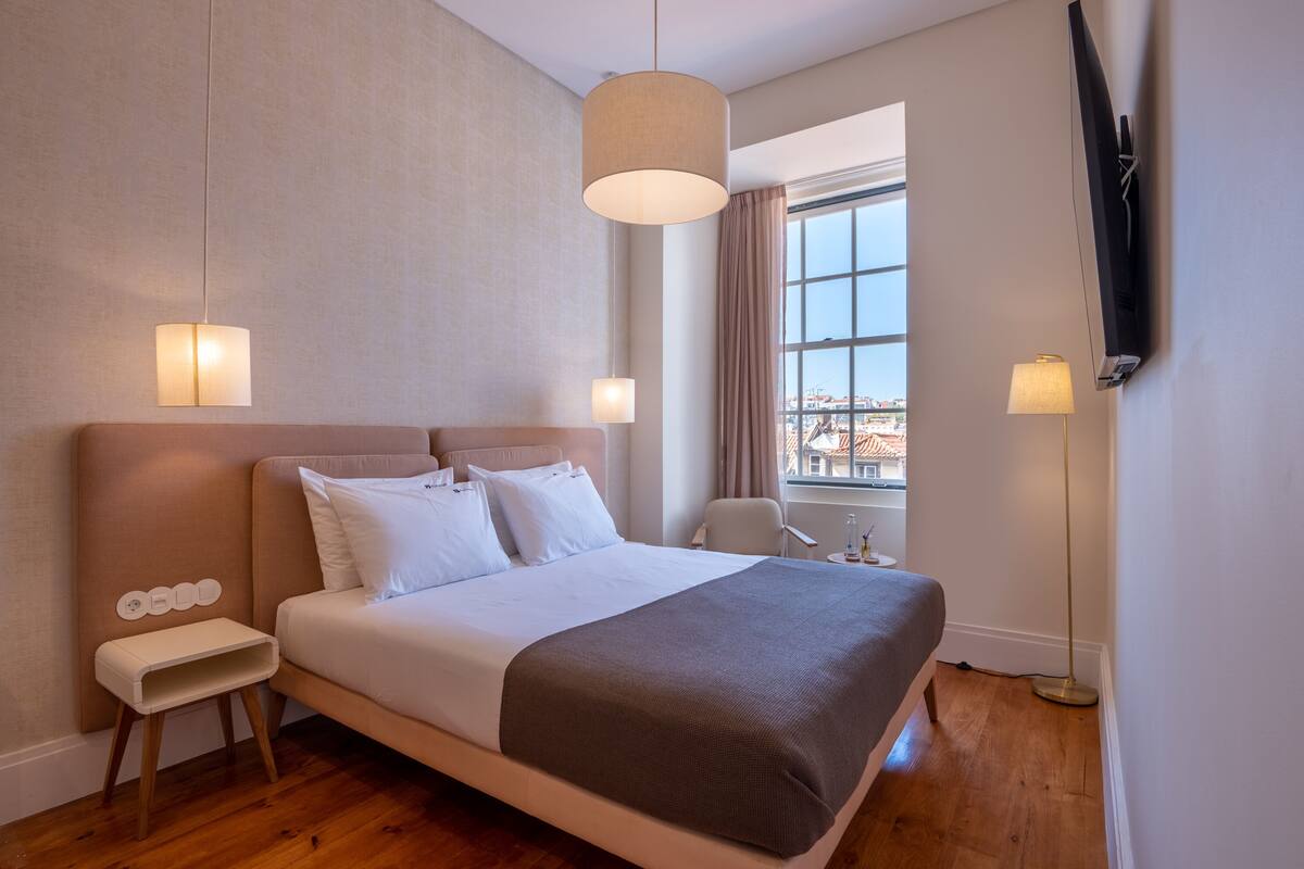 Habitación de hotel moderna con una cama grande, mesitas de noche, un televisor montado en la pared y vistas a la ciudad a través de la ventana.