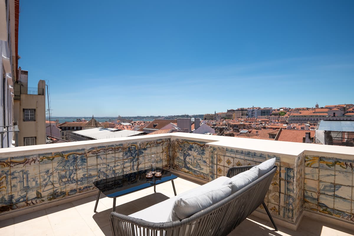 Un balcón con vistas a un paisaje urbano, una pared de azulejos con un diseño decorativo, una silla de mimbre y una pequeña mesa con un jarrón y una vela.