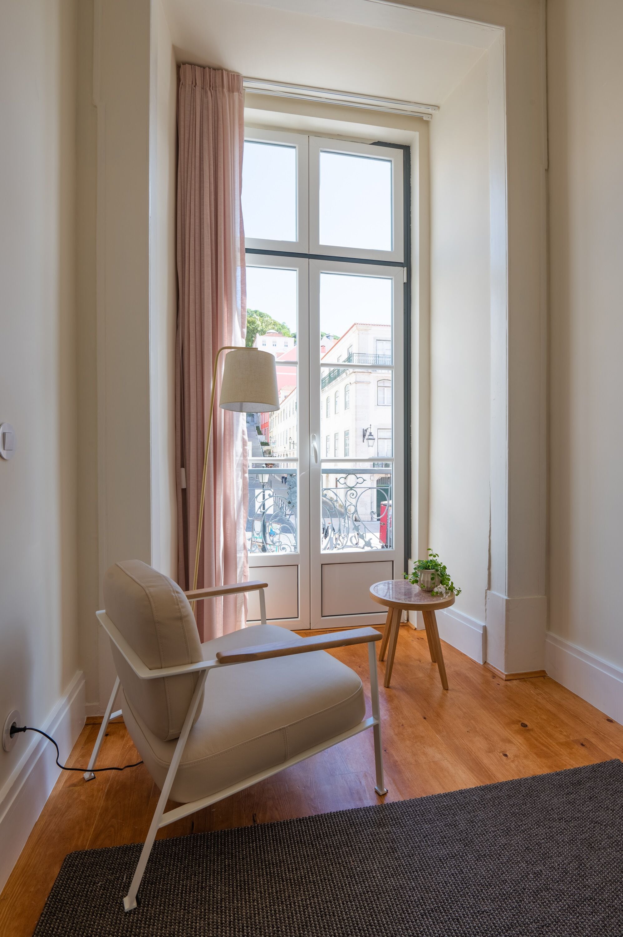 Et værelse med et stort vindue, en stol, et lille bord med en plante og en lampe.