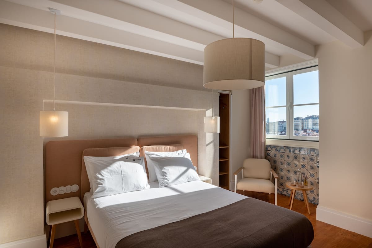 Una habitación de hotel moderna con una cama grande, una mesita de noche, una silla, un ventanal con vista a la ciudad y una lámpara colgante.