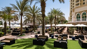 A rooftop bar with outdoor seating, palm trees, and a view of a high-rise building. - Maison Privee - Luxurious 2/Bed Apt on Palm Jumeirah (Dubai)