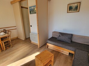A small living space with a sofa, a table, and a chair. There is a door leading to another room and a picture on the wall.