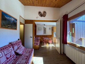 A cozy room with a wooden ceiling, a sofa with patterned cushions, a dining table with a checkered tablecloth, and a view through a window.