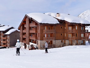 A multi-story wooden building with balconies, ski slopes in the foreground, and snow-covered mountains in the background.