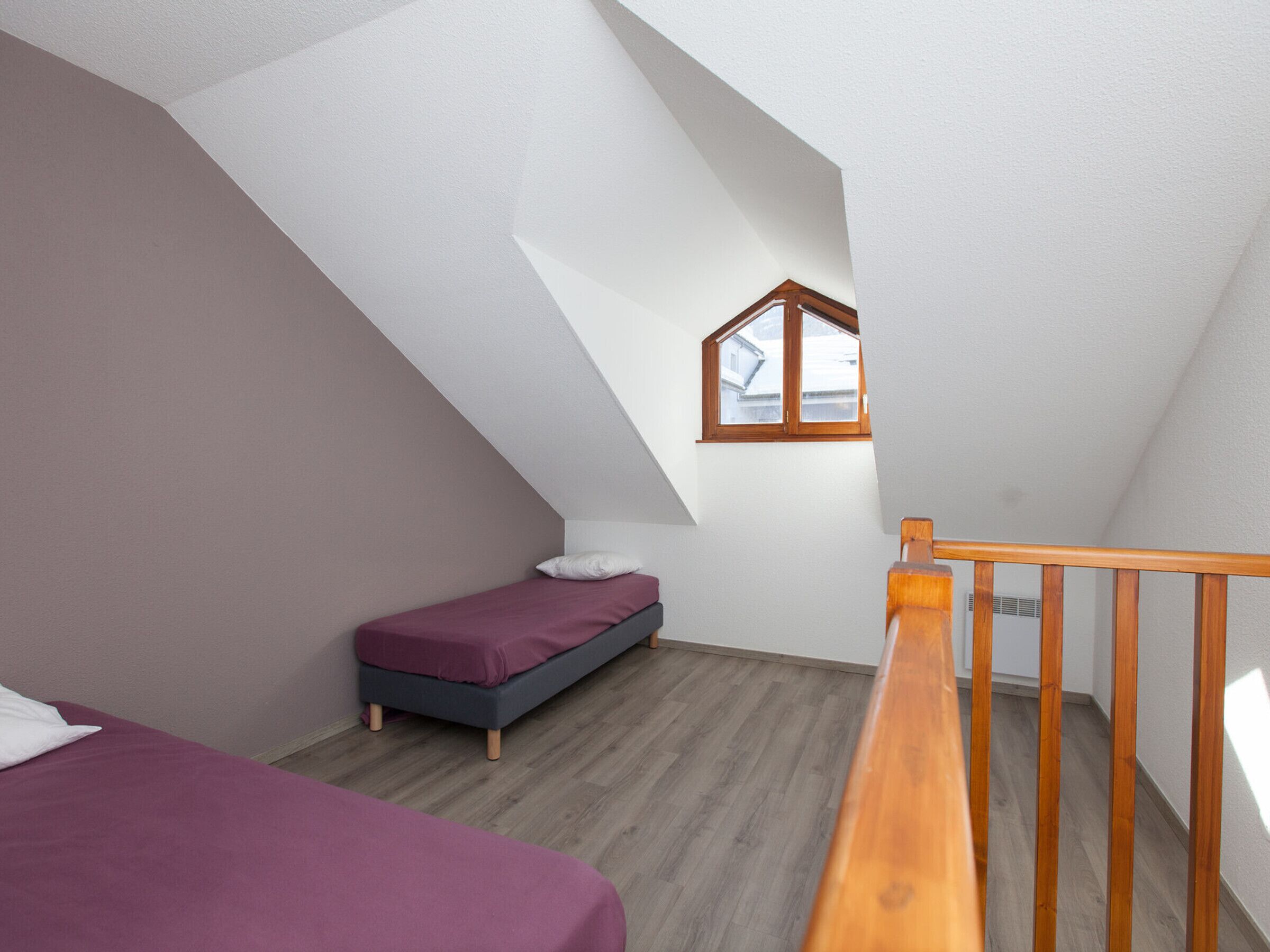 Una habitación pequeña con techo inclinado, una cama individual, baranda de madera y una ventana.