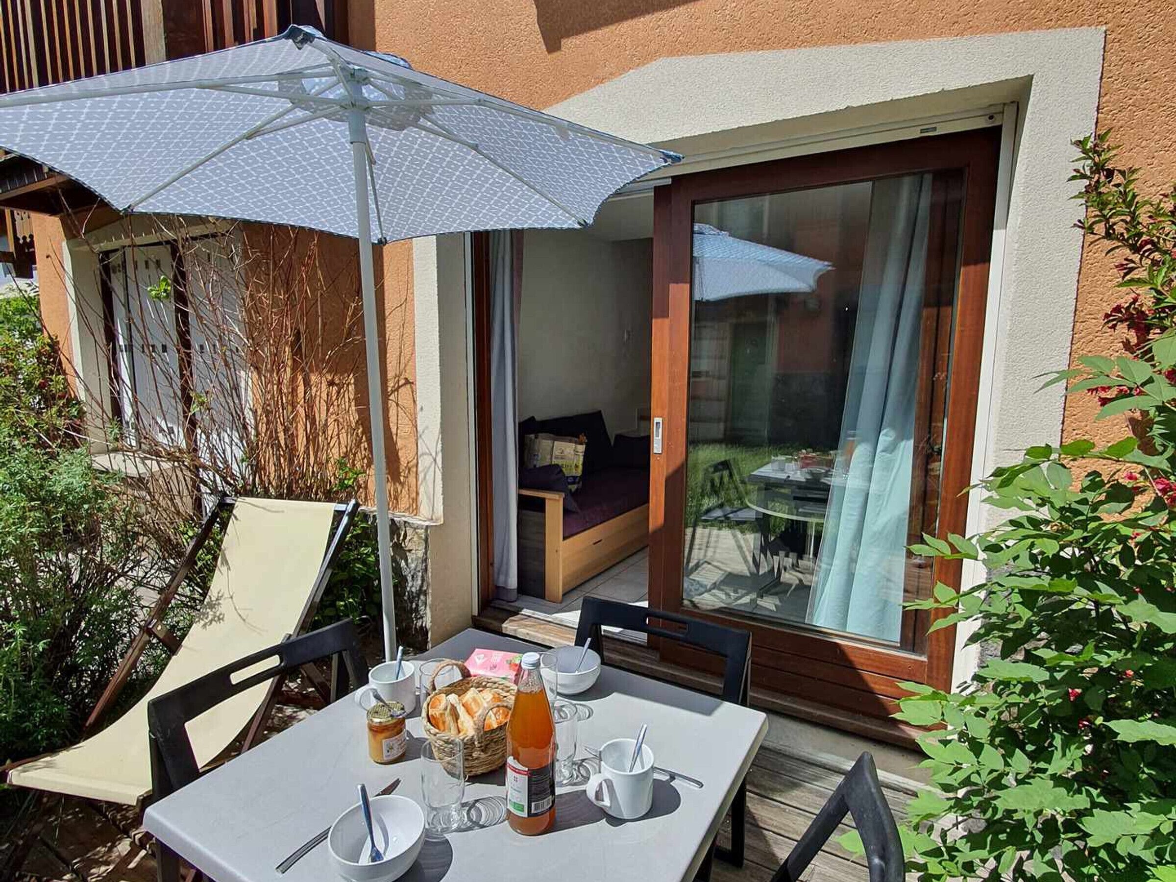 An outdoor dining area with a table set for two, a wicker basket with bread, a bottle of juice, and a glass of water.