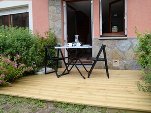 Una terraza de madera al aire libre con una mesa y sillas, rodeada de follaje y un muro de piedra.
