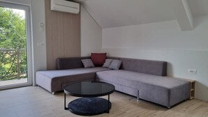 Ein modernes Wohnzimmer mit einem grauen Ecksofa, einem runden schwarzen Couchtisch und einem großen Fenster mit Blick auf Grünflächen.