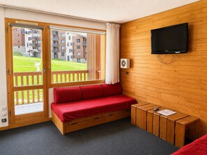 Ein Zimmer mit Holzboden, einem roten Sofa, einem an der Wand befestigten Fernseher und einem Fenster mit Blick auf Gebäude und einen Rasen.