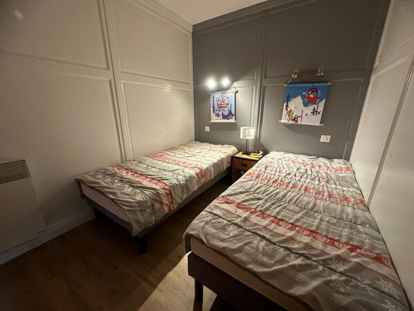 A small, well-lit room with two single beds, a nightstand with a lamp, and a wall-mounted shelf with artwork.