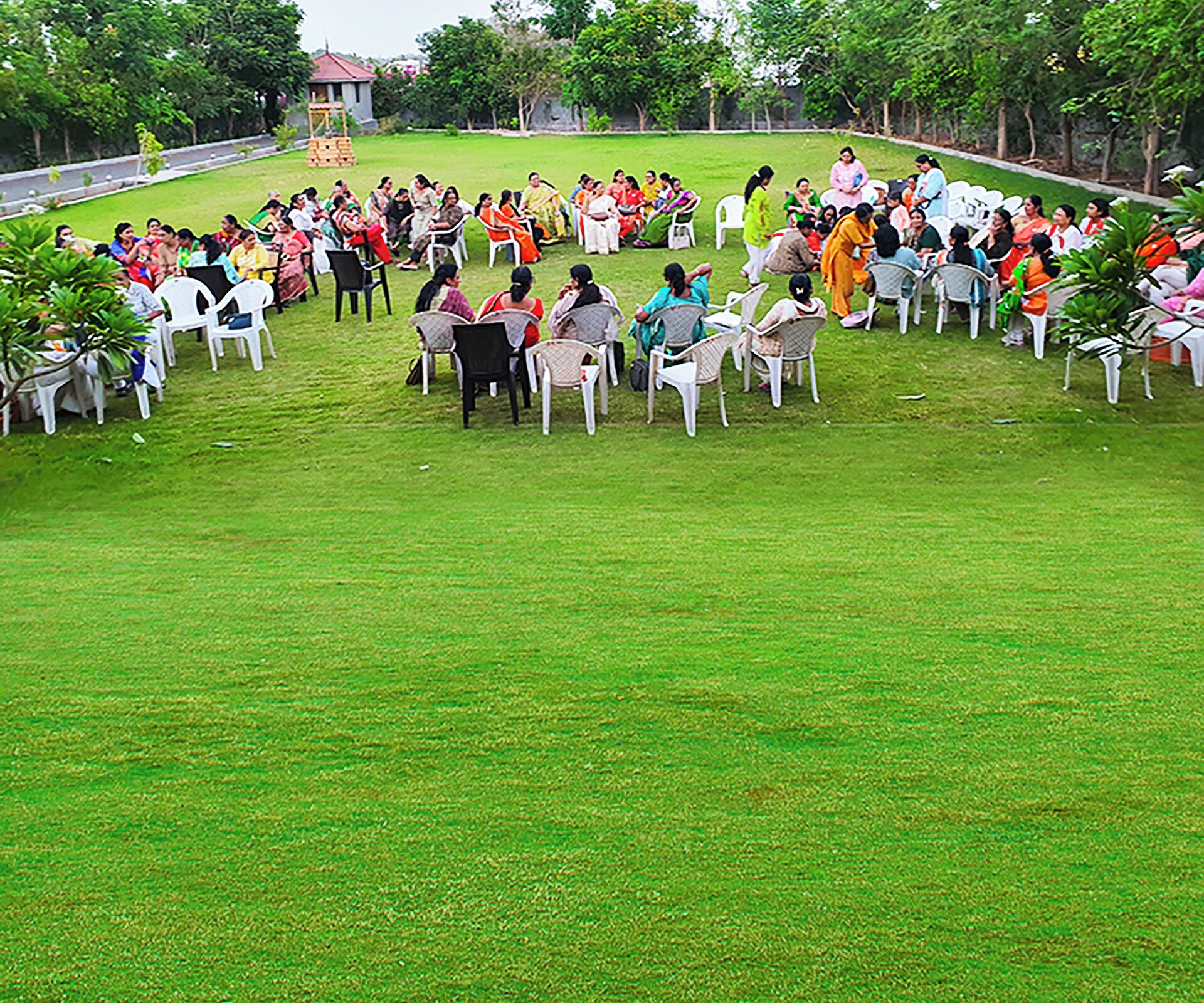 Um grupo de pessoas sentadas em círculo em um campo gramado com cadeiras brancas, cercadas por árvores.