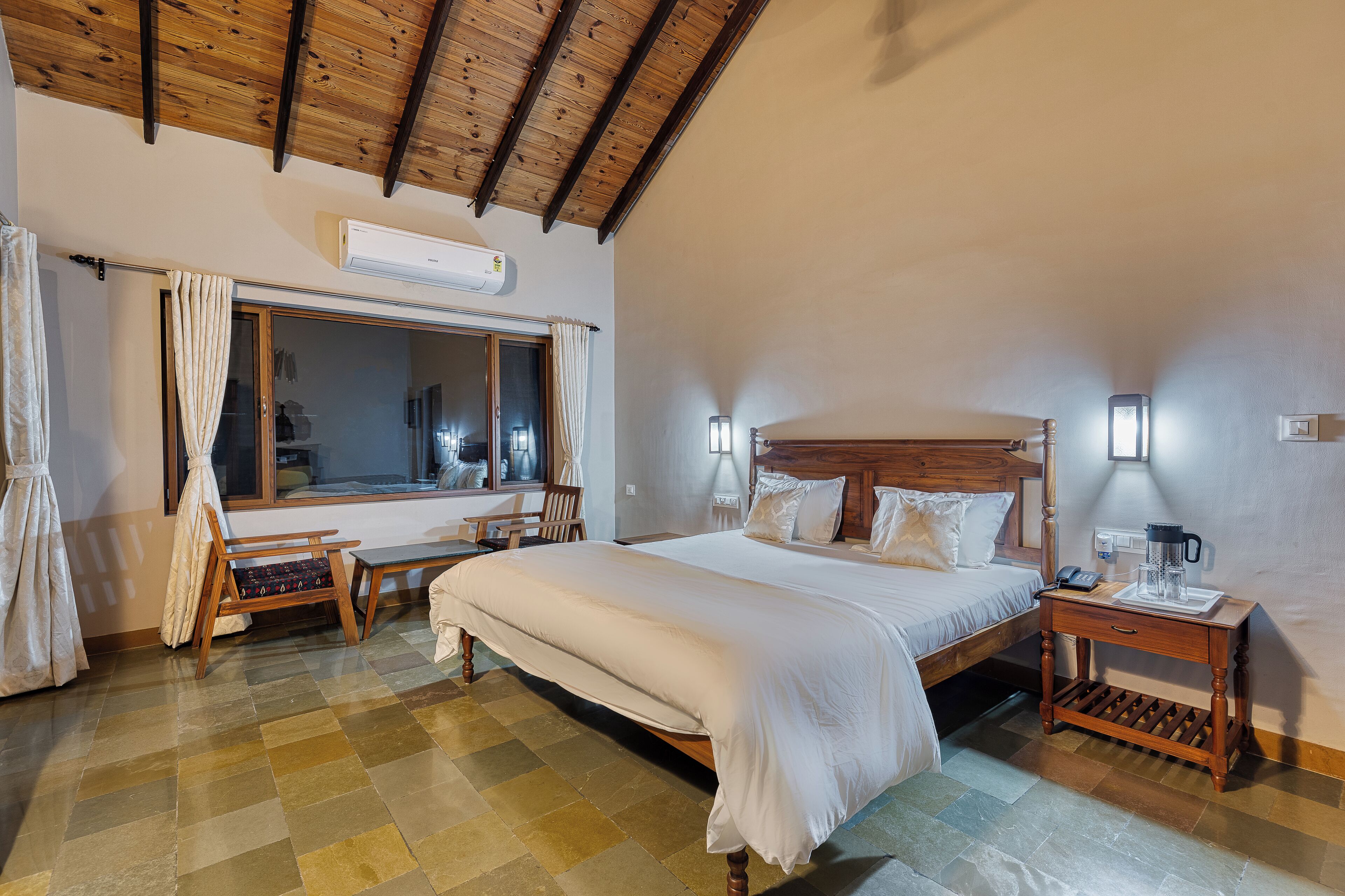 A bedroom with a large bed, wooden headboard, bedside tables, a bench, and a window with curtains.