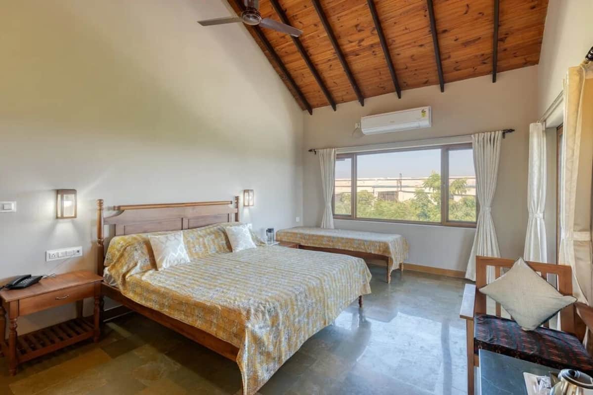 A bedroom with two beds, a wooden ceiling, a window with curtains, a bedside table with a lamp, and a chair with a cushion.