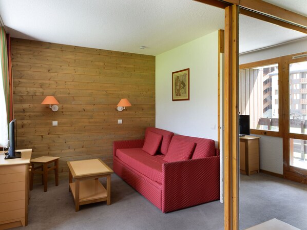 A compact room with a wooden wall, a red sofa, a small wooden table, and two wall-mounted lamps.