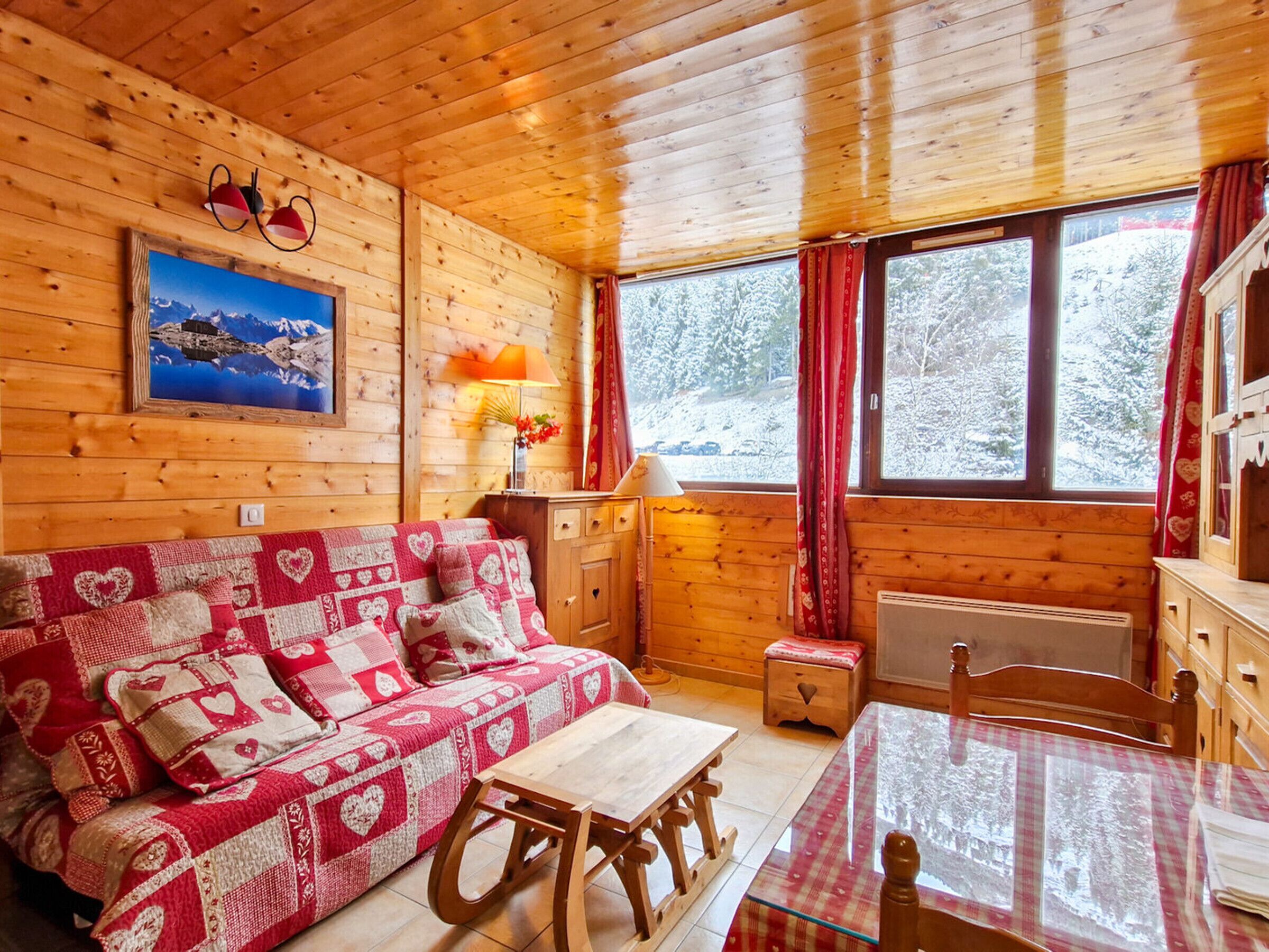 A cozy cabin interior with a wooden interior, a sofa with heart-patterned cushions, a table, and a window with a view of snow-covered trees.