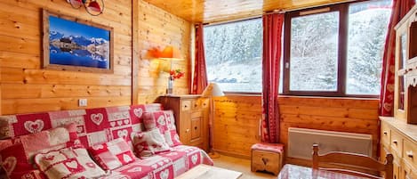 A cozy cabin interior with a wooden interior, a sofa with heart-patterned cushions, a table, and a window with a view of snow-covered trees.