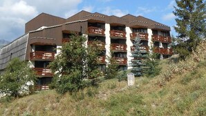 A multi-story building with balconies, surrounded by trees and a hilly landscape.