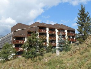 Un edificio de varios pisos con balcones, rodeado de árboles y un paisaje montañoso.