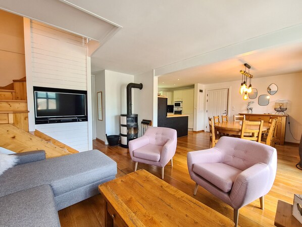A modern living room with a TV, sofa, and chairs, a kitchen area, and a staircase.