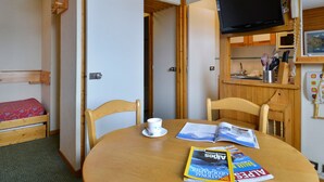 A compact room with a wooden bed, a dining table with magazines, a television, and a kitchen area with wooden cabinets and a microwave. - Functional studio at the foot of the slopes for 3/4 people, balcony, divisible living (Belle Plagne)