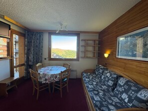 A room with a round table, two chairs, a sofa, a window with a view of a landscape, and a wall-mounted shelf.