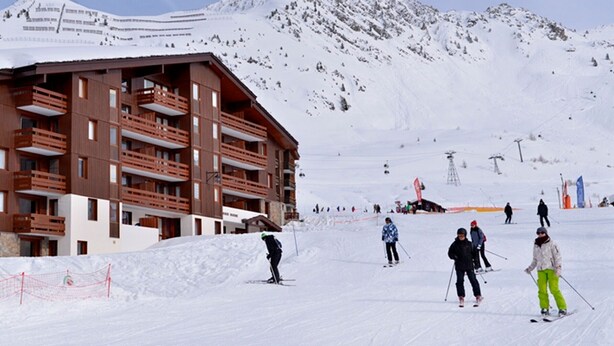 Une station de ski avec un bâtiment à plusieurs étages doté de balcons, une piste de ski et une montagne en arrière-plan.