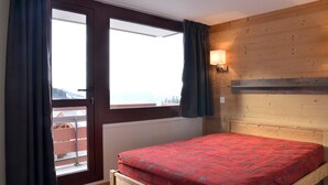 A bedroom with a wooden headboard, a bed with a red bedspread, a window with curtains, and a view of a balcony. - Renovated cozy apartment near slopes, 3 rooms + cabin for 8 people, pets allowed (Plagne Bellecôte)