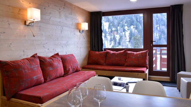 A cozy room with a wooden wall, a red sofa, a dining table with wine glasses, and a view of a forest through a window. - Renovated cozy apartment near slopes, 3 rooms + cabin for 8 people, pets allowed (Plagne Bellecôte)