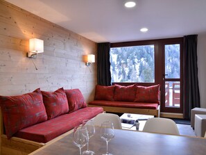 A cozy room with a wooden wall, a red sofa, a dining table with wine glasses, and a view of a forest through a window. - Renovated cozy apartment near slopes, 3 rooms + cabin for 8 people, pets allowed (Plagne Bellecôte)