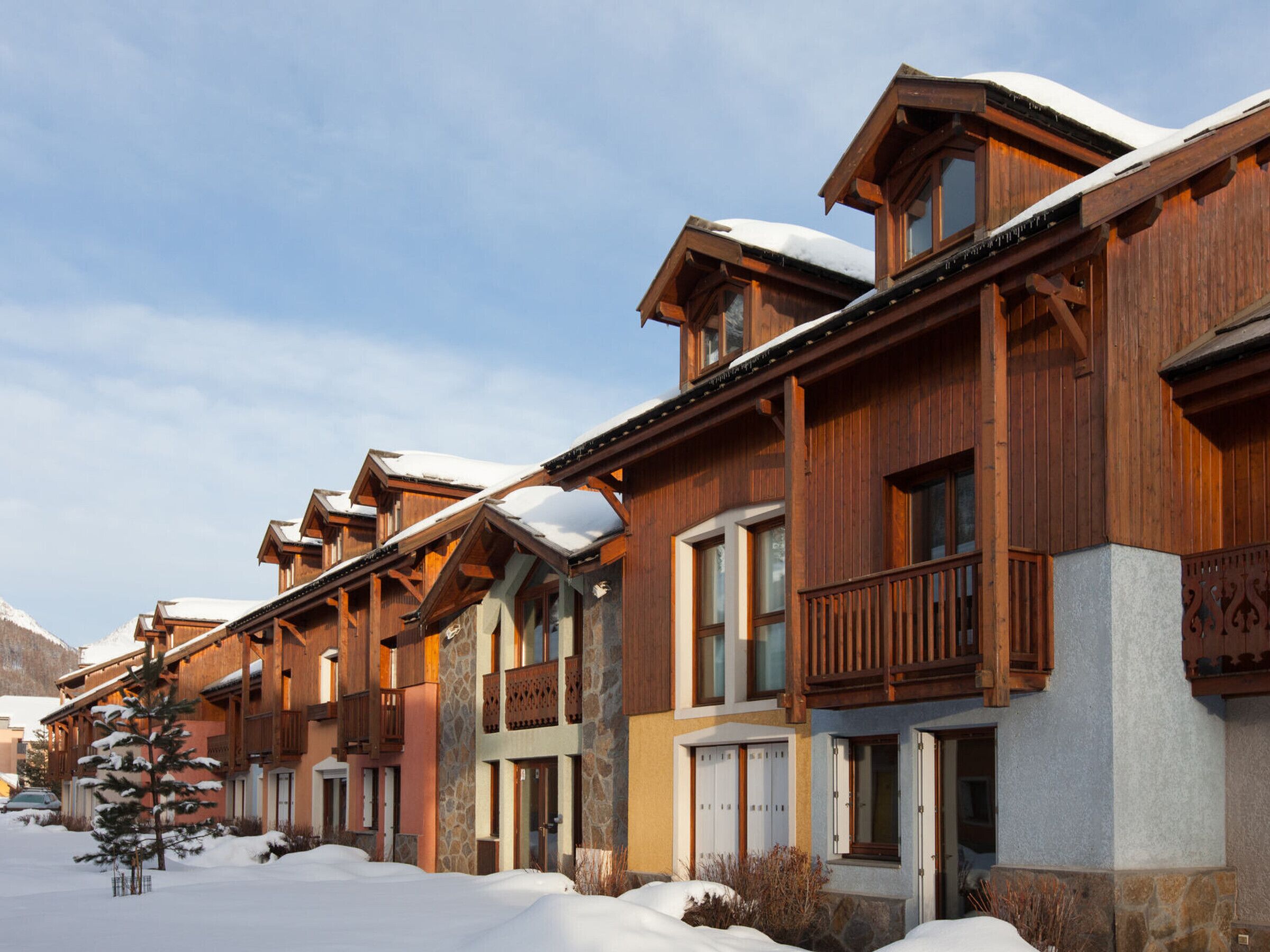 A row of wooden buildings with balconies and snow-covered roofs, likely part of a ski resort accommodation.