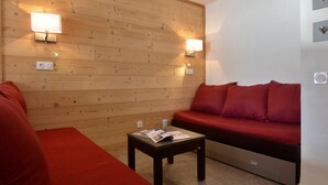 A room with a wooden wall, two wall-mounted lights, a red sofa, a small table with magazines, and a white wall with a thermostat.