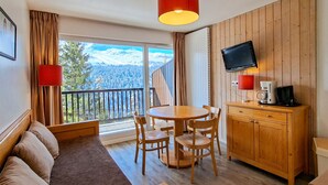 A room with a wooden floor, a round table with chairs, a sofa, a wooden cabinet with a TV, and a balcony with curtains. - Bright apartment in center with balcony and parking (Flaine)