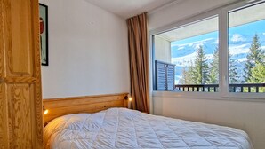 A bedroom with a wooden headboard, a white quilted bedspread, a window with a view of trees and mountains, and a curtain. - Bright apartment in center with balcony and parking (Flaine)